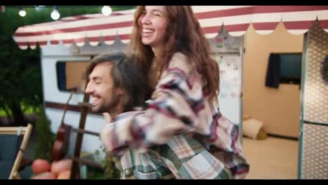 A-happy-brunette-girl-in-a-checkered-shirt-jumps-from-behind-on-her-brunette-boyfriend-in-a-green-checkered-shirt,-and-he-picks-her-up-and-begins-to-spin-her-during-his-vacation-outside-the-city-in-a-camp-near-a-trailer-in-the-summer