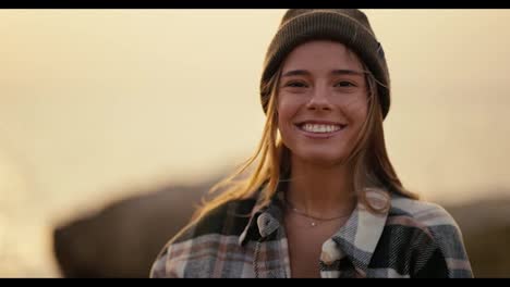 Portrait-of-a-happy-blonde-girl-in-a-black-hat-in-a-plaid-shirt-who-looks-at-the-camera-and-smiles-while-standing-on-a-rocky-shore-near-the-sea