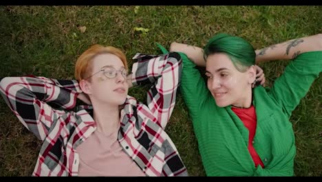 Close-up-shot-of-a-blonde-girl-with-a-short-haircut-in-glasses-with-sparkles-on-her-face-in-a-checkered-shirt-and-her-girlfriend-with-a-short-green-haircut-in-a-green-shirt-are-talking-Lesha-on-the-green-grass-in-the-park