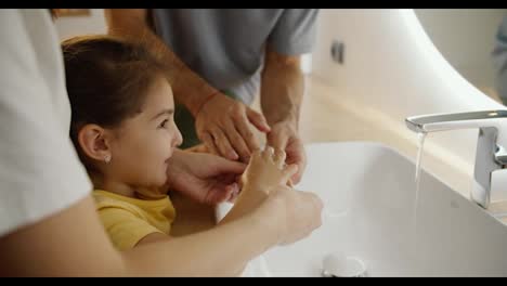 Close-up-shot-of-a-little-brunette-girl-in-a-white-T-shirt-washes-her-hands-and-is-helped-in-this-by-her-dad-and-mom-near-a-modern-sink-and-a-round-illuminated-mirror-in-the-bathroom