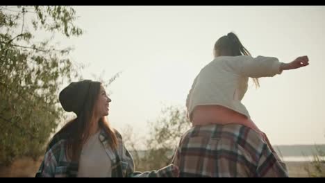 A-little-blonde-girl-in-a-white-jacket-sits-on-the-shoulders-of-her-dad,-the-brunette-in-a-brown-checkered-shirt,-and-next-to-her-is-her-mother,-the-brunette-girl-in-a-green-checkered-shirt,-they-have-fun-together,-communicate-and-travel-outside-the-city