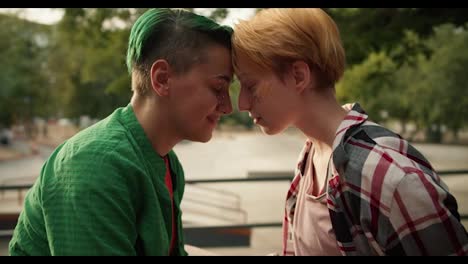 Two-lesbian-girls,-a-girl-with-a-green-haircut-and-green-hair-in-a-green-shirt-and-a-girl-with-yellow-hair-in-a-plaid-shirt,-lean-their-faces-against-each-other-and-rub-their-noses-during-their-date-at-a-skate-park-in-the-summer