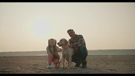 Portrait-of-a-happy-middle-aged-man-with-gray-hair-in-a-plaid-shirt-who-sits-near-his-large-cream-colored-dog-and-his-little-blonde-girl-daughter-in-a-white-jacket-and-pink-pants-on-a-deserted-seashore-during-his-vacation-outside-the-city-in-a-summer-evening