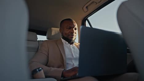 A-confident-brunette-man-with-Black-skin-in-a-brown-suit-works-on-a-gray-laptop-during-his-business-trip-outside-the-urban-area