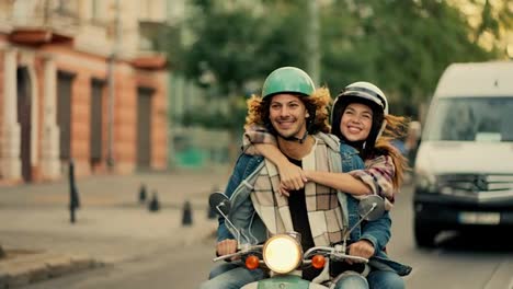 Happy-couple-guy-with-long-curly-hair-and-in-a-denim-jacket-rides-with-his-happy-girlfriend-in-a-plaid-shirt-who-holds-tightly-to-her-boyfriend-smiling-broadly-and-looking-at-him-while-riding-a-moped-on-a-city-summer-street