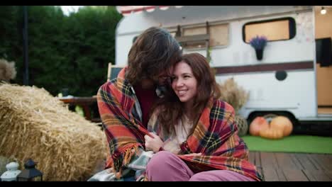 Happy-brunette-guy-in-a-checkered-plaid-sits-wrapped-up-with-his-brunette-girlfriend-during-his-picnic-near-a-trailer-outside-the-city-in-a-camp-in-the-summer