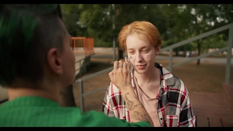 Over-the-shoulder:-A-girl-with-a-short-haircut-with-green-hair-in-a-green-shirt-applies-glitter-with-her-finger-to-the-face-of-her-girlfriend-with-short-orange-hair-in-a-checkered-pink-shirt-on-brown-benches-in-the-park