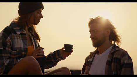 Ein-Blondes-Mädchen-Mit-Schwarzem-Hut-Und-Kariertem-Hemd-Gibt-Ihrem-Freund-Bei-Sonnenaufgang-In-Der-Nähe-Des-Meeres-Eine-Tasse-Heißen-Tee-Aus-Einer-Thermoskanne