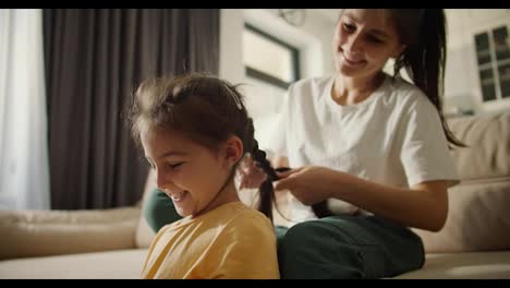 Happy-brunette-mother-in-a-white-T-shirt-gives-her-cheerful-brunette-daughter-in-a-yellow-dress-a-braid-hairstyle-while-sitting-on-a-light-brown