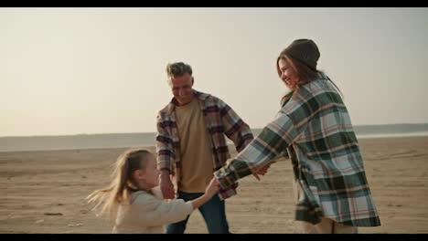 A-happy-trio-of-a-family,-a-brunette-man-in-a-checkered-shirt,-together-with-his-brunette-wife-in-a-green-checkered-shirt-and-a-little-girl-daughter-in-a-white-jacket,-dance-on-a-deserted-seashore-and-spin-in-a-circle-and-dance-in-a-circle-during-their-picnic-and-vacation-outside-the-city