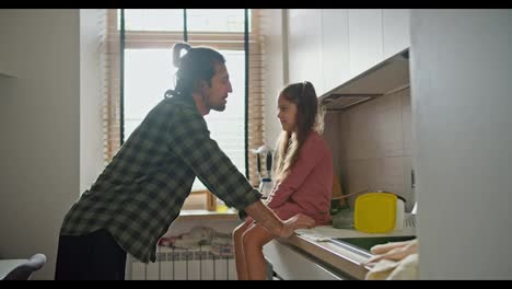 A-brunette-man-in-a-green-checkered-shirt-leans-on-the-table-and-communicates-with-his-little-brunette-daughter-in-a-pink-dress-in-a-modern-kitchen