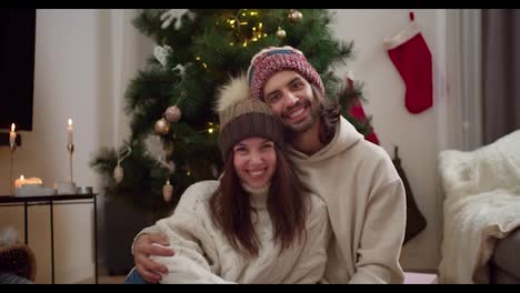 Portrait-of-a-happy-couple,-a-guy-and-a-girl-in-warm-New-Year's-hats-and-white-sweaters-are-sitting-next-to-each-other,-smiling-and-looking-at-the-camera-near-the-New-Year's-tree-in-a-cozy-apartment-in-winter