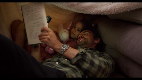 A-brunette-man-in-a-green-checkered-shirt-together-with-his-little-daughter,-a-brunette-girl-in-a-pink-dress-who-is-holding-a-unicorn-toy-in-her-hands,-reading-a-book-under-a-makeshift-awning-at-home-near-a-gray-sofa