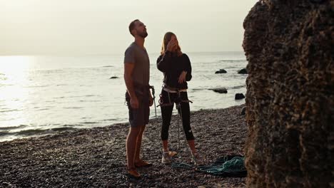 A-blonde-girl-with-curly-hair-in-a-black-Sweater-tells-a-guy-in-a-gray-T-shirt-how-they-will-climb-a-rock-near-a-rocky-seashore-and-then-insures-the-guy-so-that-he-climbs-up