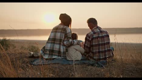 Rear-view-of-a-happy-brunette-girl-in-a-green-checkered-shirt-along-with-her-middle-aged-man-husband-in-a-checkered-shirt-and-little-daughter-sitting-on-a-mat-outside-the-city-and-watching-the-sunset-during-their-picnic-in-the-summer