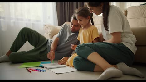 Der-Glückliche-Junge-Familienvater-Zeichnet-Zusammen-Mit-Seiner-Tochter-Und-Seiner-Frau-Mit-Bunten-Bleistiften-Auf-Papier,-Während-Er-In-Einem-Modernen-Zimmer-Auf-Dem-Boden-Neben-Dem-Sofa-Sitzt