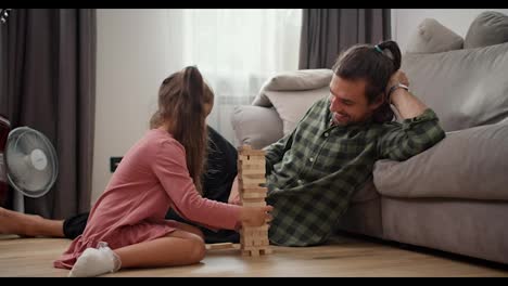 Vista-Lateral-De-Una-Pequeña-Niña-Morena-Con-Un-Vestido-Rosa-Juega-Con-Su-Padre-Moreno-Con-Una-Camisa-Verde-A-Cuadros-El-Juego-De-Mesa-Jenga-Y-Cuando-Saca-Uno-De-Los-Ladrillos-La-Torre-Jenga-Se-Cae