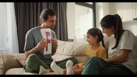 A-brunette-man-in-a-gray-T-shirt-teaches-his-little-daughter-in-a-yellow-dress-various-letters,-showing-them-on-pieces-of-paper.-Little-girl's-parents-help-her-learn-different-letters-by-showing-colorful-letters-on-a-sheet-of-paper