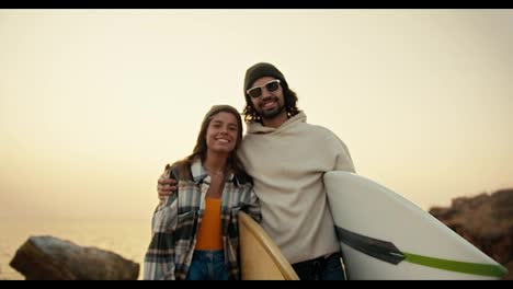 Portrait-of-a-happy-couple,-a-brunette-man-in-a-white-sweatshirt-and-black-sunglasses-together-with-his-blonde-girlfriend-in-a-plaid-shirt-stand-near-the-sea-and-hold-surfboards-in-their-hands-in-the-morning-in-autumn