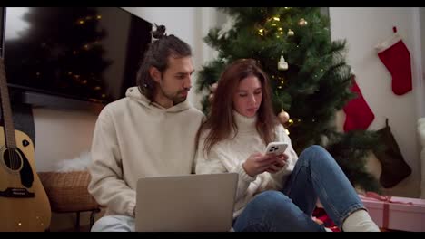 Serious-and-thoughtful-brunette-guy-and-girl-in-white-sweaters-sit-on-the-floor-and-work-on-a-laptop-and-phone-near-the-New-Year-tree-in-a-cozy-room-in-a-winter-evening