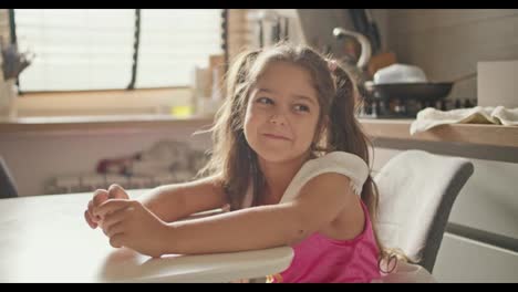 Portrait-of-a-happy-little-brunette-girl-in-a-pink-dress-who-sits-at-the-kitchen-table-in-a-modern-kitchen-during-the-day.-A-little-girl-in-a-fairy-dress-sits-at-the-table-in-the-kitchen,-rejoices-and-has-fun