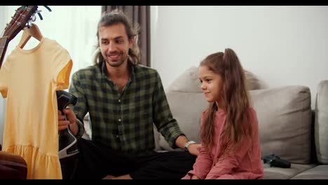 A-brunette-man-with-long-hair-wearing-a-green-plaid-shirt-together-with-his-brunette-daughter,-a-little-girl-in-a-pink-jacket-steaming-a-yellow-T-shirt-using-a-special-steamer-while-sitting-on-a-gray-sofa-in-a-modern-apartment