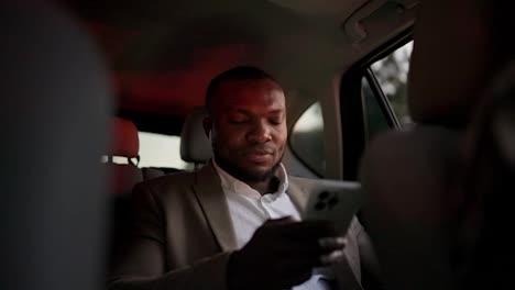 A-confident-man-with-Black-skin-in-a-brown-suit-works-on-his-white-smartphone-while-driving-on-a-business-trip-in-the-interior-of-a-modern-car