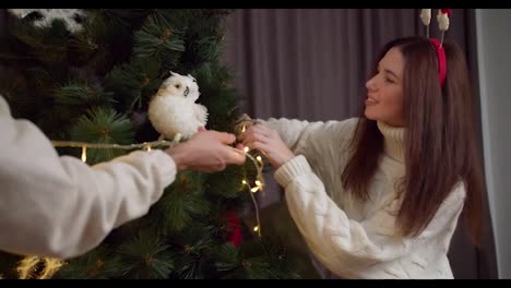 Close-up-shot-of-a-brunette-girl-in-a-white-sweater,-together-with-her-boyfriend,-decorate-the-New-Year-tree-and-with-the-help-of-a-bright-yellow-glowing-garland-and-prepare-for-Christmas-in-winter-at-home