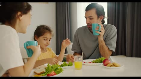 A-brunette-man-in-a-gray-T-shirt-holds-a-turquoise-cup-in-his-hand-and-has-breakfast-with-his-wife-and-little-daughter-in-a-yellow-dress-with-a-white-table-in-a-modern-kitchen