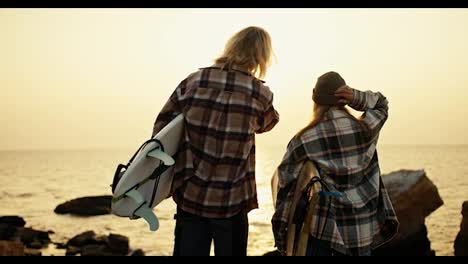 Rear-view-of-a-tall-blond-man-in-a-plaid-shirt-and-his-blond-girlfriend-in-a-hat-and-plaid-shirt-are-holding-surfboards-while-standing-on-a-rocky-shore-near-the-sea-and-looking-at-the-rising-Sun-in-the-morning-in-summer