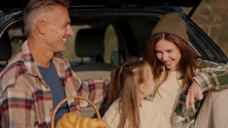 Happy-brunette-girl-in-a-hat-in-a-green-checkered-shirt-communicates-with-her-husband,-a-middle-aged-man-in-a-checkered-shirt,-and-her-little-daughter,-a-blonde-girl,-during-their-picnic-outside-the-city-near-the-car-in-the-summer