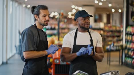 A-man-with-Black-skin-in-a-white-T-shirt-and-a-black-apron-in-blue-protective-rubber-gloves-together-with-his-colleague-a-brunette-man-with-a-beard-communicate-and-discuss-their-work-plan-in-a-large-supermarket