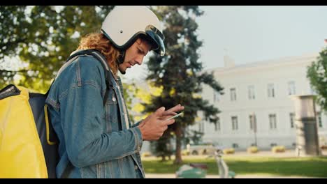 A-courier-guy-with-curly-hair-in-a-denim-shirt-walks-through-a-summer-park,-looks-around-and-looks-at-his-phone-for-navigation-and-gets-on-his-moped-with-a-large-yellow-bag-and-leaves-to-deliver-his-order-in-the-summer-city