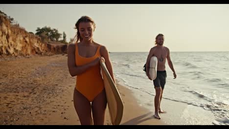 Una-Chica-Rubia-Feliz-Y-Segura-De-Sí-Misma-Con-Un-Traje-De-Baño-Naranja-Camina-Por-La-Orilla-Rocosa-Y-Arenosa-Del-Mar-Y-Lleva-Una-Tabla-De-Surf-Detrás-De-Ella,-Su-Novio-Rubio-Con-Pantalones-Negros-Camina-Y-También-Lleva-Una-Tabla-De-Surf-Cerca-Del-Mar-En-La-Orilla-Por-La-Mañana.