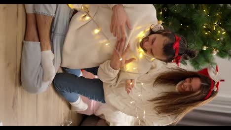 Vertical-video,-a-brunette-girl-in-a-White-sweater-decorates-her-brunette-boyfriend-in-a-red-New-Year's-hoop-with-the-help-of-a-yellow-glowing-garland-near-a-green-New-Year's-tree-in-a-cozy-room