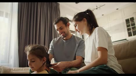 Solo-Un-Hombre-Moreno-Junto-Con-Su-Novia-Morena-Con-Una-Camiseta-Blanca-Están-Aprendiendo-A-Hacer-Un-Peinado-Trenzado-Para-Su-Pequeña-Hija-Con-Un-Vestido-Amarillo-Sentada-Sobre-Un-Marrón-Claro-En-Un-Acogedor-Apartamento-Moderno.