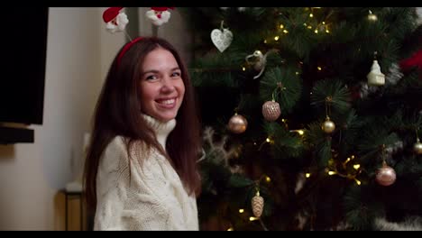 Portrait-of-a-happy-young-brunette-girl-in-a-white-sweater-who-smiles-and-looks-at-the-camera-near-her-decorated-Christmas-tree-in-a-cozy-winter-home