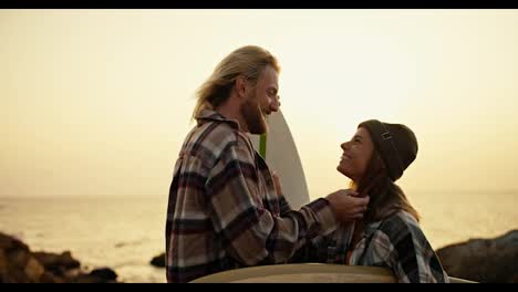 Happy-blond-guy-with-a-beard-in-a-checkered-shirt-kisses-his-blonde-girlfriend,-who-holds-a-surfboard-and-rejoices-on-a-rocky-shore-near-the-sea.-Romantic-meeting-of-surfers,-guy-and-girl-on-a-date-in-the-morning