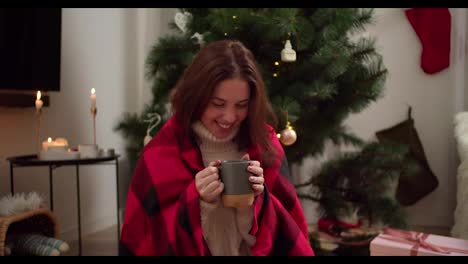 Happy-brunette-girl-in-a-red-checkered-plaid-drinks-hot-tea-from-a-gray-mug,-smiles-and-looks-to-the-side-near-the-Christmas-Tree-in-a-cozy-room-in-the-evening-in-winter