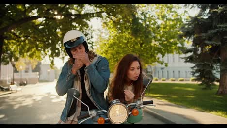 A-brunette-girl-preens-herself-looking-into-the-side-mirror-of-a-moped,-and-her-boyfriend-in-a-denim-shirt-in-a-White-helmet-pretentiously-smokes-a-cigarette-and-looks-at-his-girlfriend-in-a-sunny-summer-city-on-the-street