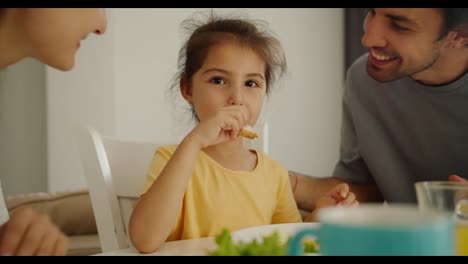 Portrait-of-a-happy-little-brunette-girl-in-a-yellow-dress-who-is-having-breakfast-and-her-two-parents-kiss-her-on-both-cheeks-during-breakfast-at-the-family-table-in-the-morning-in-a-modern-apartment