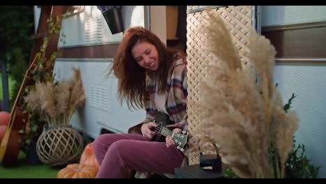 Happy-brunette-girl-in-a-checkered-shirt-and-pink-jeans-plays-a-black-ukulele-near-her-trailer-outside-the-city-in-the-summer-during-a-picnic.-Yellow-sprigs-of-reeds-in-a-vase-and-other-decorations-of-the-trailer-during-their-day-off-outside-the-city-in-the-summer-on-a-picnic