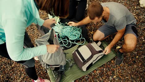 Shooting-close-up-from-above,-a-guy-and-a-girl-are-disassembling-their-backpacks-and-taking-out-a-rope-and-other-equipment-for-rock-climbing-on-a-rocky-seashore-near-them-their-dog