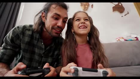 Happy-male-father-in-a-green-checkered-shirt-plays-with-his-daughter,-a-little-brunette-girl-in-a-pink-dress,-using-joysticks,-they-have-fun-and-rejoice-while-sitting-on-a-gray-sofa-at-home-in-a-modern-apartment