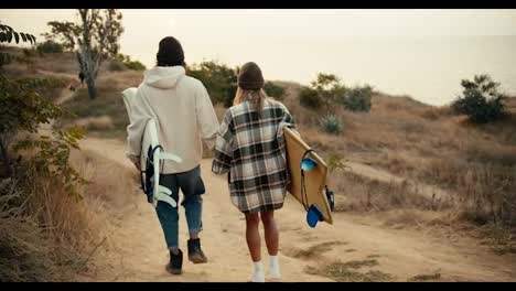 Rear-view-of-a-happy-couple,-a-brunette-guy-in-a-hat-in-a-white-sweatshirt-and-a-blonde-girl-in-a-hat-in-a-checkered-shirt-walk-down-the-slope-and-carry-boards-for-surfing-near-the-sea-in-the-summer