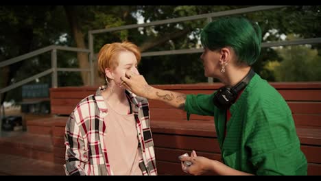 A-girl-with-green-hair-in-a-green-shirt-with-headphones-makes-makeup-for-her-girlfriend-with-bright-orange-hair-in-a-plaid-shirt-on-brown-seats-near-the-skate-park-in-the-summer-in-the-park