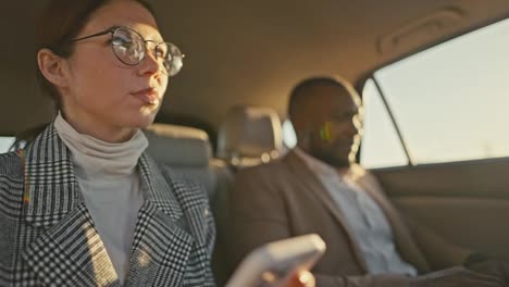 A-confident-girl-businesswoman-in-round-glasses-in-a-gray-jacket-chats-on-her-smartphone-during-her-trip-and-business-trip-together-with-a-male-businessman-with-Black-skin-in-a-brown-jacket-in-a-modern-salon-in-a-car-on-the-road