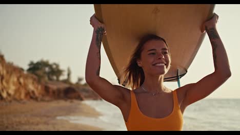 A-happy-and-cheerful-blonde-girl-in-an-orange-swimsuit-carries-her-light-wooden-surfboard-above-her-head-and-walks-along-the-sandy-rocky-seashore-in-the-morning-at-Sunrise