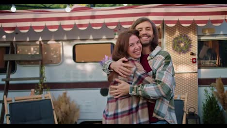 Portrait-of-a-happy-brunette-girl-in-a-pink-checkered-shirt-who-runs-to-her-brunette-boyfriend-in-a-green-checkered-shirt-and-hugs-him-in-their-trailer-while-relaxing-in-a-camp-during-a-picnic-in-the-summer