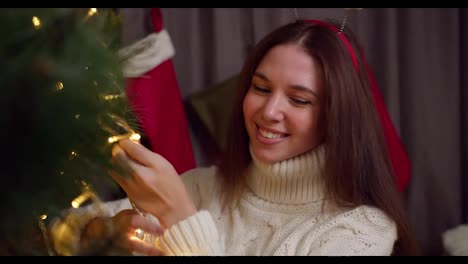 Close-up-shot-of-a-happy-brunette-girl-in-a-white-sweater-decorates-the-New-Year-tree-with-the-help-of-a-yellow-glowing-garland-in-a-cozy-home-in-winter-and-prepares-for-the-New-Year-holidays-and-Christmas